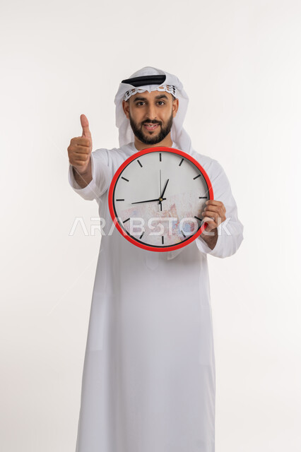 التذكيرات والتنبيهات الثابته، الدقة في المواعيد، بورتريه لرجل عربي خليجي اماراتي يرتدي الكندورة والغترة ينظر الى الكاميرا يحمل بيده ساعة رقمية كبيرة يرفع اصبع الابهام بعلامة الاعجاب والرضا, إنجاز المهامات في الوقت، خلفية بيضاء
