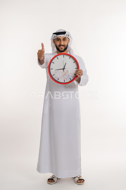 الدقة في المواعيد، بورتريه لرجل عربي خليجي اماراتي يرتدي الكندورة والغترة ينظر الى الكاميرا يحمل بيده ساعة رقمية كبيرة يرفع اصبع الابهام بعلامة الاعجاب والرضا, التذكيرات والتنبيهات الثابته، إنجاز المهامات في الوقت، صورة الجسم الطول الكامل، خلفية بيضاء