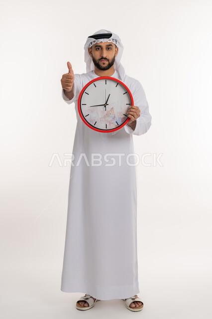الدقة في المواعيد، بورتريه لرجل عربي خليجي اماراتي يرتدي الكندورة والغترة ينظر الى الكاميرا يحمل بيده ساعة رقمية كبيرة يرفع اصبع الابهام بعلامة الاعجاب والرضا, التذكيرات والتنبيهات الثابته، إنجاز المهامات في الوقت، صورة الجسم الطول الكامل، خلفية بيضاء