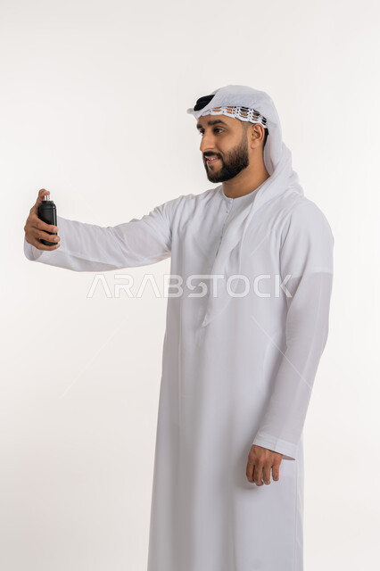 Perfuming with luxurious perfumes and scents, using natural essential oils, a portrait of an Arab Gulf Emirati man wearing a kandora and ghutra, holding a bottle in his hand, spraying perfume, the concept of elegance and concern for external appearance, white background