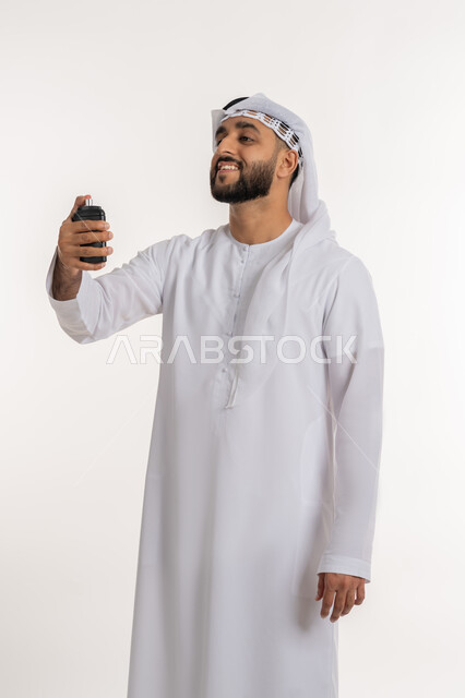 التطيب بالعطور والروائح الفاخرة، استخدام الزيوت العطرية الطبيعية، بورتريه لرجل عربي خليجي اماراتي يرتدي الكندورة والغترة يمسك بيده زجاجة يقوم برش العطر, مفهوم الأناقة والاهتمام بالمظهر الخارجي، خلفية بيضاء