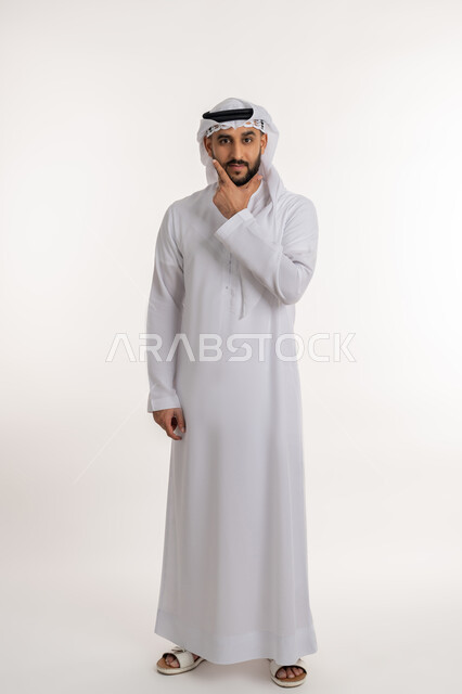 التمعن والتركيز لإيجاد حل لمشكلة، بورتريه لرجل عربي خليجي اماراتي يرتدي الكندورة والغترة يضع يده على ذقنه بإيماءات التفكير بشيء ما, التأمل و التخطيط للمستقبل، صورة الجسم بالطول الكامل، خلفية بيضاء