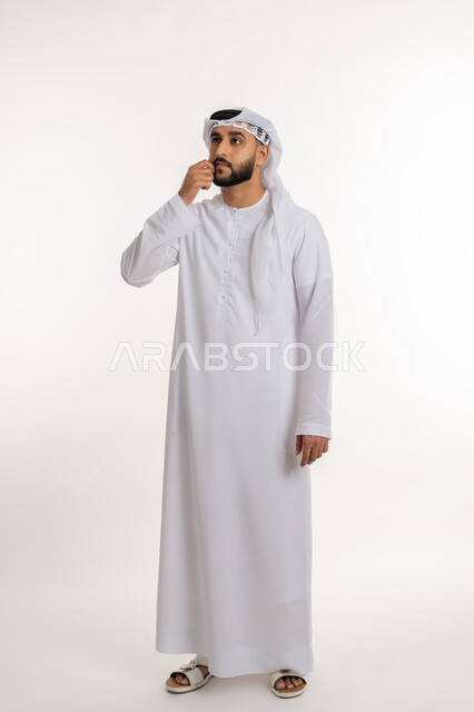 التمعن والتركيز لإيجاد حل لمشكلة، بورتريه لرجل عربي خليجي اماراتي يرتدي الكندورة والغترة يضع يده على ذقنه بإيماءات التفكير بشيء ما, التأمل و التخطيط للمستقبل، صورة الجسم بالطول الكامل، خلفية بيضاء