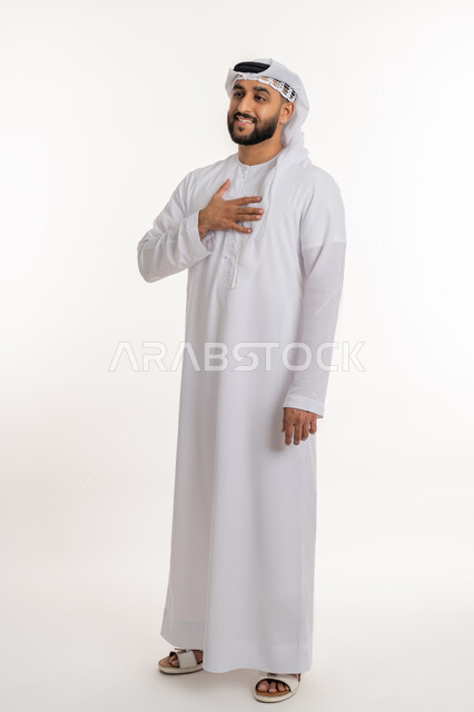 مفهوم رحابة الصدر والشكر والامتنان، بورتريه لرجل عربي خليجي اماراتي يرتدي الكندورة والغترة يضع يده على صدره بإيماءات الترحيب والاستقبال, تعابير السرور والفرح، صورة الجسم بالطول الكامل, خلفية بيضاء