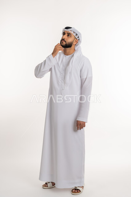 التأمل و التخطيط للمستقبل، التمعن والتركيز لإيجاد حل لمشكلة، بورتريه لرجل عربي خليجي اماراتي يرتدي الكندورة والغترة يضع يده على ذقنه بإيماءات التفكير بشيء ما, صورة الجسم بالطول الكامل, خلفية بيضاء