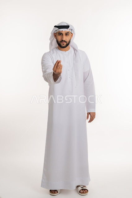 إمهال بعض الوقت للقيام بشئ ما، مفهوم الصبر والتريث، بورتريه لرجل عربي خليجي اماراتي يرتدي الكندورة والغترة ينظر الى الكاميرا يشير بيده بإيماءات طلب الإنتظار، صورة الجسم بالطول الكامل، خلفية بيضاء