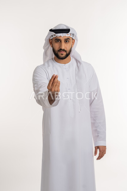 إمهال بعض الوقت للقيام بشئ ما، مفهوم الصبر والتريث، بورتريه لرجل عربي خليجي اماراتي يرتدي الكندورة والغترة ينظر الى الكاميرا يشير بيده بإيماءات طلب الإنتظار، خلفية بيضاء