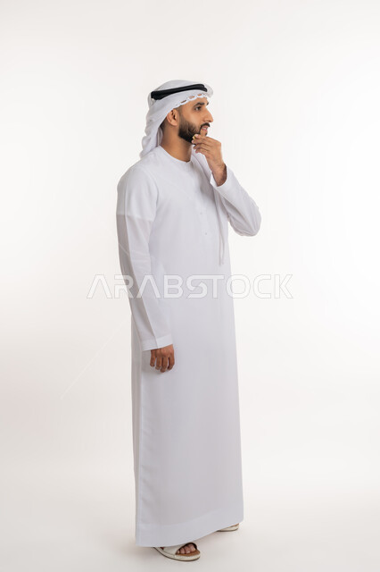 Focusing and contemplating to find a solution to a problem, portrait of an Arab Gulf Emirati man wearing a kandora and ghutra, placing his hand on his chin with gestures of thinking about something, contemplating and planning for the future, full-length body portrait, white background