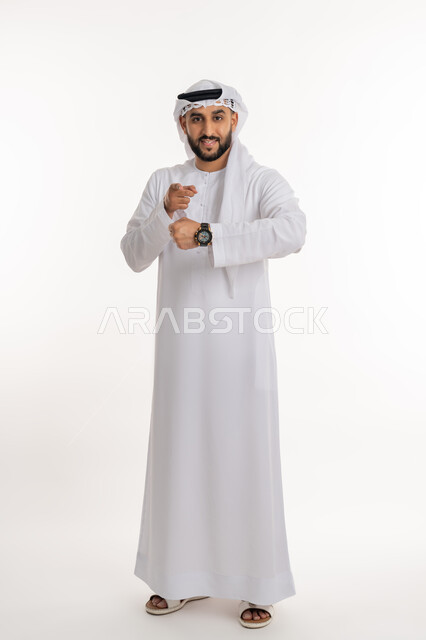 تنظيم ومراقبة الوقت، تحديد مواعيد العمل، إدارة الجداول الزمنية، بورتريه لرجل عربي خليجي اماراتي يرتدي الكندورة والغترة يرتدي ساعة اليد ينظر الى الكاميرا يشير باصبع السبابة، صورة الجسم بالطول الكامل, خلفية بيضاء