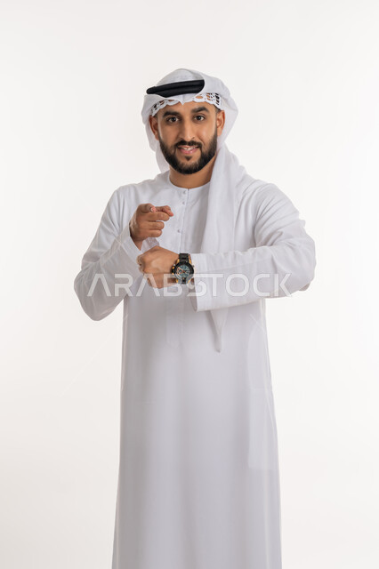 تنظيم ومراقبة الوقت، تحديد مواعيد العمل، إدارة الجداول الزمنية، بورتريه لرجل عربي خليجي اماراتي يرتدي الكندورة والغترة يرتدي ساعة اليد ينظر الى الكاميرا يشير باصبع السبابة، خلفية بيضاء