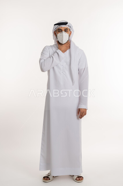 صعوبة التنفس والتعب والألم، العدوى بفايروس كورونا، بورتريه لرجل عربي خليجي اماراتي يرتدي الكندورة والغترة والكمامة الواقية ينظر الى الكاميرا يضع يديه على حلقه يشعر بالاختناق، الاصابة بنزلة برد والتهاب الحلق، صورة الجسم بالطول الكامل, خلفية بيضاء