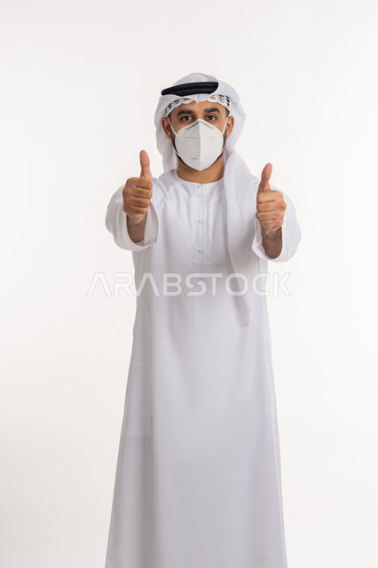 تجنب انتشار العدوى بكوفيد 19، النظر الى الكاميرا بتعابير الحذر، بورتريه لرجل عربي خليجي اماراتي يرتدي الكندورة والغترة والكمامة الواقية يرفع اصبع الابهام للاعلى بإيماءات القبول والرضا، الالتزام بإجراءات الوقاية، خلفية بيضاء