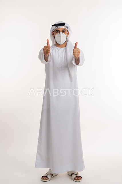 تجنب انتشار العدوى بكوفيد 19، بورتريه لرجل عربي خليجي اماراتي يرتدي الكندورة والغترة والكمامة الواقية ينظر الى الكاميرا يرفع اصبع الابهام للاعلى بإيماءات القبول والرضا، الالتزام بإجراءات الوقاية، صورة الجسم بالطول الكامل، خلفية بيضاء