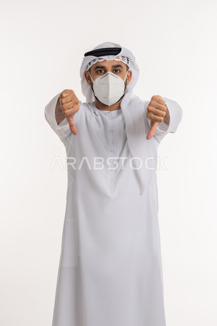تجنب انتشار العدوى بكوفيد 19، بورتريه لرجل عربي خليجي اماراتي يرتدي الكندورة والغترة والكمامة الواقية ينظر الى الكاميرا يرفع اصبع الابهام للاسفل بإيماءات الرفض وعدم القبول، الالتزام بإجراءات الوقاية، خلفية بيضاء
