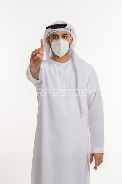 الالتزام بإجراءات الوقاية، تجنب انتشار العدوى بكوفيد 19، بورتريه لرجل عربي خليجي اماراتي يرتدي الكندورة والغترة والكمامة الواقية ينظر الى الكاميرا يرفع اصبع السبابة للاعلى بإيماءات التحذير والتنبيه، خلفية بيضاء