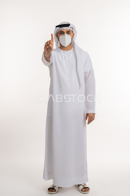 الالتزام بإجراءات الوقاية، تجنب انتشار العدوى بكوفيد 19، بورتريه لرجل عربي خليجي اماراتي يرتدي الكندورة والغترة والكمامة الواقية ينظر الى الكاميرا يرفع اصبع السبابة للاعلى بإيماءات التحذير والتنبيه، صورة الجسم بالطول الكامل, خلفية بيضاء