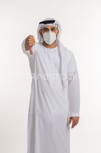 تجنب انتشار العدوى بكوفيد 19، بورتريه لرجل عربي خليجي اماراتي يرتدي الكندورة والغترة والكمامة الواقية ينظر الى الكاميرا يرفع اصبع الابهام للاسفل بإيماءات الرفض وعدم القبول، الالتزام بإجراءات الوقاية، خلفية بيضاء