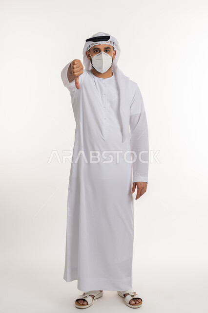 تجنب انتشار العدوى بكوفيد 19، بورتريه لرجل عربي خليجي اماراتي يرتدي الكندورة والغترة والكمامة الواقية ينظر الى الكاميرا يرفع اصبع الابهام للاسفل بإيماءات الرفض وعدم القبول، الالتزام بإجراءات الوقاية، صورة الجسم بالطول الكامل, خلفية بيضاء