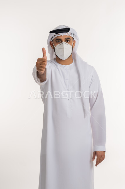 تجنب انتشار العدوى بكوفيد 19، النظر الى الكاميرا بتعابير الحذر، بورتريه لرجل عربي خليجي اماراتي يرتدي الكندورة والغترة والكمامة الواقية يرفع اصبع الابهام للاعلى بإيماءات القبول والرضا، الالتزام بإجراءات الوقاية، خلفية بيضاء