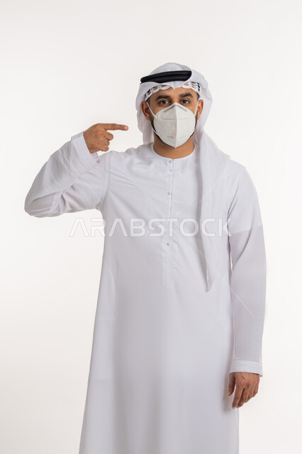 Looking at the camera with an expression of caution, a portrait of an Arab Gulf Emirati wearing a kandora, ghutra, and a protective mask, pointing with his index finger to the mask, adhering to prevention and safety measures, avoiding the spread of infection with Covid-19, white background