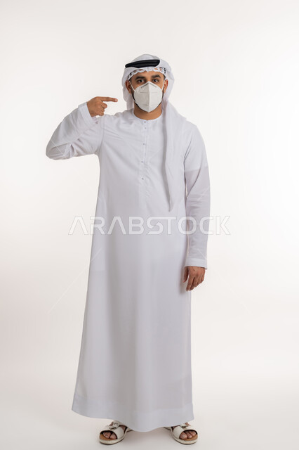 النظر الى الكاميرا بتعابير الحذر، بورتريه لرجل عربي خليجي اماراتي يرتدي الكندورة والغترة والكمامة الواقية يشير بإصبع السبابة الى القناع، الالتزام بإجراءات الوقاية والسلامة، تجنب انتشار العدوى بكوفيد 19، صورة الجسم بالطول الكامل, خلفية بيضاء