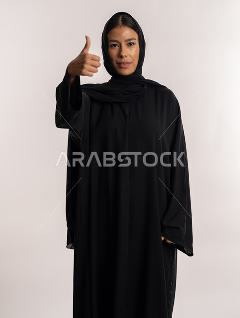 تعابير القبول و الموافقة, الإشارة إلى الجودة و الامتياز, بورتريه لامرأة عربية خليجية إماراتية ترتدي العباءة السوداء تنظر إلى الكاميرا بإيماءات الإعجاب و الرضا, شابة سعودية تشير بإصبع الإبهام للأعلى, خلفية بيضاء