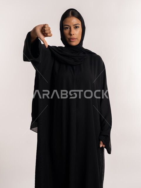 تعابير الجدية و الحزم, حركات الرفض و عدم الموافقة, بورتريه لامرأة عربية خليجية إماراتية ترتدي العباءة السوداء تنظر إلى الكاميرا بإيماءات عدم الرضا, شابة سعودية تشير بإصبع الإبهام للأسفل, خلفية بيضاء