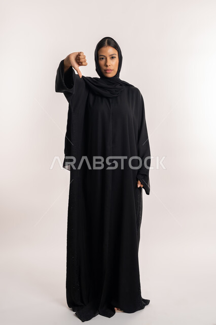 تعابير الجدية و الحزم, حركات الرفض و عدم الموافقة, بورتريه لامرأة عربية خليجية إماراتية ترتدي العباءة السوداء تنظر إلى الكاميرا بإيماءات عدم الرضا, شابة سعودية تشير بإصبع الإبهام للأسفل, صورة للجسم بالطول الكامل, خلفية بيضاء