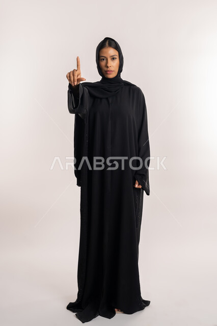 لفت الانتباه إلى أمر مهم, تعابير التحذير و التأكيد على نقطة معينة, بورتريه مقرب لامرأة عربية خليجية إماراتية ترتدي العباءة السوداء تنظر إلى الكاميرا بجدية, شابة سعودية تشير بإصبع السبابة إلى الأعلى بإيماءات التوجيه, الجسم بالطول الكامل, خلفية بيضاء