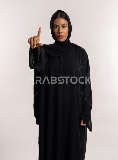 تعابير التحذير و التأكيد على نقطة معينة, لفت الانتباه إلى أمر مهم, بورتريه لامرأة عربية خليجية إماراتية ترتدي العباءة السوداء تنظر إلى الكاميرا بتعابير جدية, شابة سعودية تشير بإصبع السبابة إلى الأعلى بإيماءات التوجيه, خلفية بيضاء