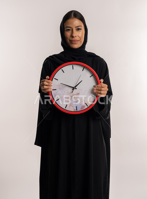 التذكيرات و التنبيهات الثابتة, طرق تنظيم و إدارة الوقت, بورتريه لامرأة عربية خليجية إماراتية ترتدي العباءة السوداء تنظر إلى الكاميرا بإيماءات السرور, شابة سعودية تمسك ساعة الحائط الحمراء بيدها, خلفية بيضاء