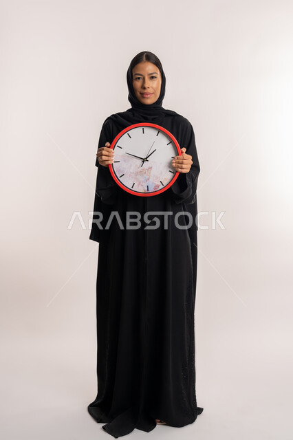 طرق تنظيم و إدارة الوقت, التذكيرات و التنبيهات الثابتة, بورتريه لامرأة عربية خليجية إماراتية ترتدي العباءة السوداء تنظر إلى الكاميرا بإيماءات السرور, شابة سعودية تمسك ساعة الحائط الحمراء بيدها, الجسم بالطول الكامل, خلفية بيضاء