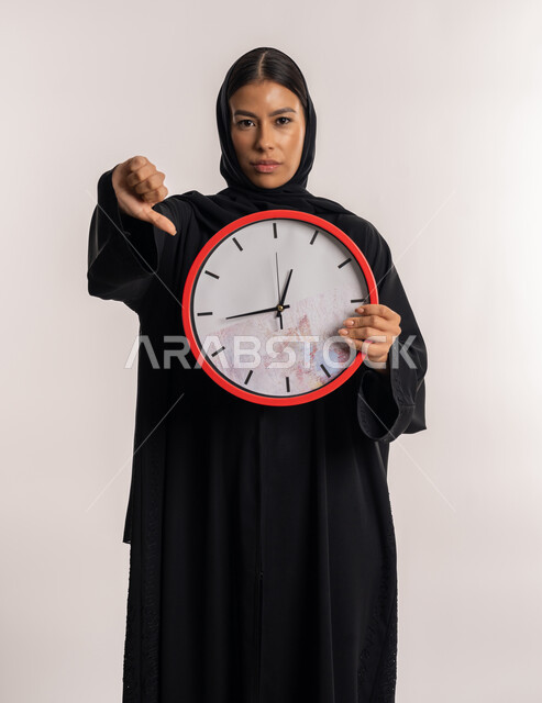 تعابير الرفض و عدم الموافقة, التذكيرات و التنبيهات الثابتة, بورتريه لامرأة عربية خليجية إماراتية ترتدي العباءة السوداء تمسك ساعة الحائط الحمراء بيدها, شابة سعودية تشير بإصبع الإبهام للأسفل, خلفية بيضاء