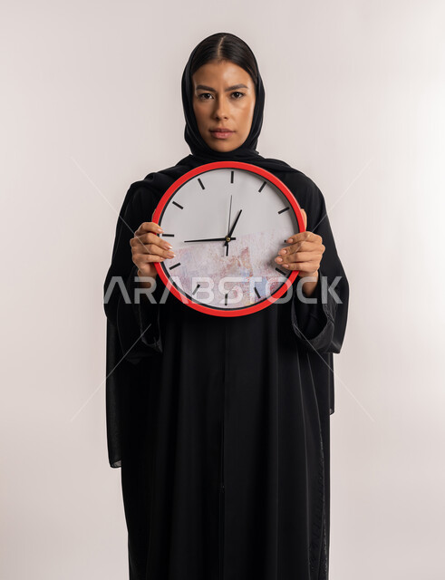 مفهوم الدقة في المواعيد, إنجاز المهام في الموعد المحدد, بورتريه لامرأة عربية خليجية إماراتية ترتدي العباءة السوداء تنظر إلى الكاميرا بتعابير الجدية, شابة سعودية تمسك بيدها ساعة حائط حمراء, خلفية بيضاء