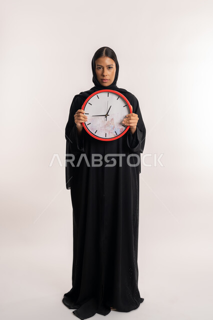 مفهوم الدقة في المواعيد, إنجاز المهام في الموعد المحدد, بورتريه لامرأة عربية خليجية إماراتية ترتدي العباءة السوداء تنظر إلى الكاميرا بتعابير الجدية, شابة سعودية تمسك بيدها ساعة حائط حمراء, الجسم بالطول الكامل, خلفية بيضاء