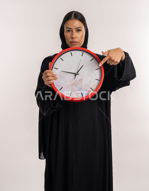 Completing tasks on time, constant reminders and alerts, the importance of keeping times, portrait of an Emirati Gulf Arab woman wearing a black abaya holding a red wall clock in her hand, a young Saudi woman pointing with her index finger to a specific time, white background
