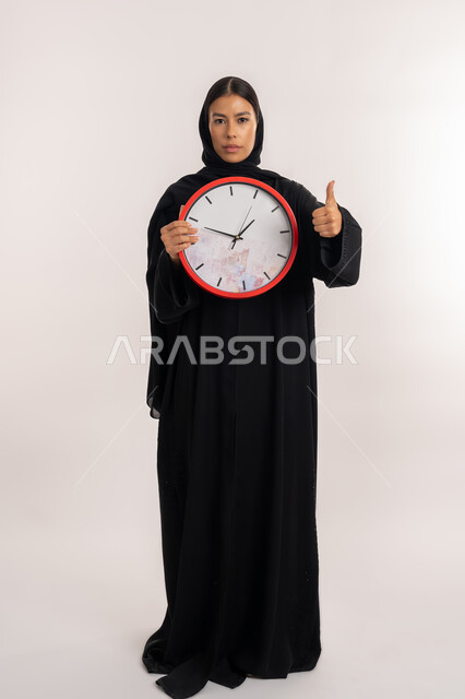 مفهوم الدقة في المواعيد, طرق تنظيم و إدارة الوقت, بورتريه لامرأة عربية خليجية إماراتية ترتدي العباءة السوداء تمسك بيدها ساعة حائط حمراء, شابة سعودية تشير بإصبع الإبهام للأعلى بتعابير الرضا و القبول, صورة للجسم بالطول الكامل, خلفية بيضاء