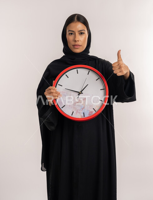 Methods of organizing and managing time, the concept of punctuality, a portrait of an Emirati Gulf Arab woman wearing a black abaya holding a red wall clock in her hand, a young Saudi woman pointing with her thumb upward with an expression of satisfaction and acceptance, a white background
