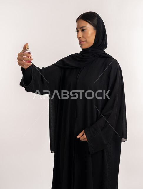 The concept of perfuming and perfuming, marketing of Gulf and international women’s perfume products, a portrait of an Arab Gulf Emirati woman wearing a black abaya, holding a perfume bottle in her hand and looking at it, a young Saudi woman using perfume, white background