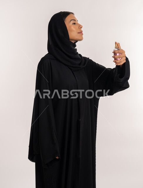 التسويق لمنتجات العطور و المستحضرات النسائية, مفهوم التطيب و التعطر, بورتريه لامرأة عربية خليجية إماراتية ترتدي العباءة السوداء تمسك زجاجة عطرية بيدها, شابة سعودية تقوم برش العطر, خلفية بيضاء