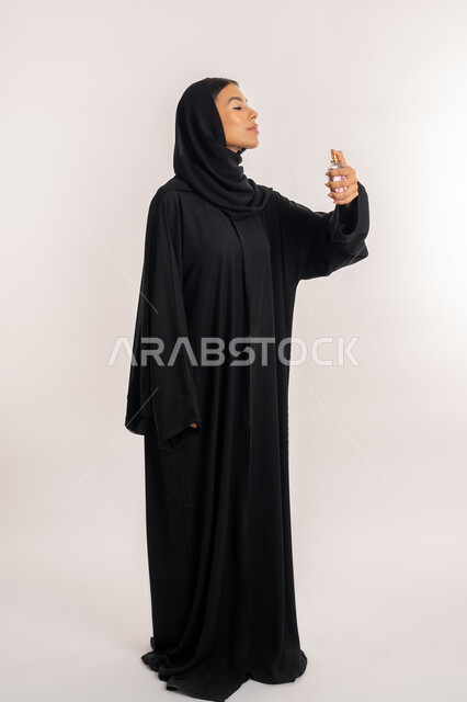 مفهوم التطيب و التعطر, التسويق لمنتجات العطور و المستحضرات النسائية, بورتريه لامرأة عربية خليجية إماراتية ترتدي العباءة السوداء تمسك زجاجة عطرية بيدها, شابة سعودية تقوم برش العطر, صورة للجسم بالطول الكامل, خلفية بيضاء