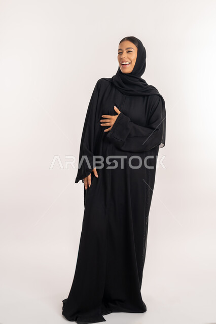 الفرح عند سماع خبر معين, بورتريه لامرأة مبتسمة عربية خليجية إماراتية ترتدي العباءة السوداء تغمض عينيها بتعابير الضحك الهستيري, شابة سعودية تضع يدها على صدرها بإيماءات السعادة و السرور, صورة للجسم بالطول الكامل, خلفية بيضاء