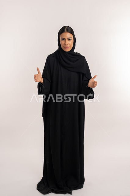 التردد و انعدام المعرفة بخصوص شيء معين, بورتريه لامرأة عربية خليجية إماراتية ترتدي العباءة السوداء تنظر إلى الكاميرا بجدية, شابة سعودية تفتح ذراعيها بإيماءات الغضب, صورة للجسم بالطول الكامل, خلفية رمادية