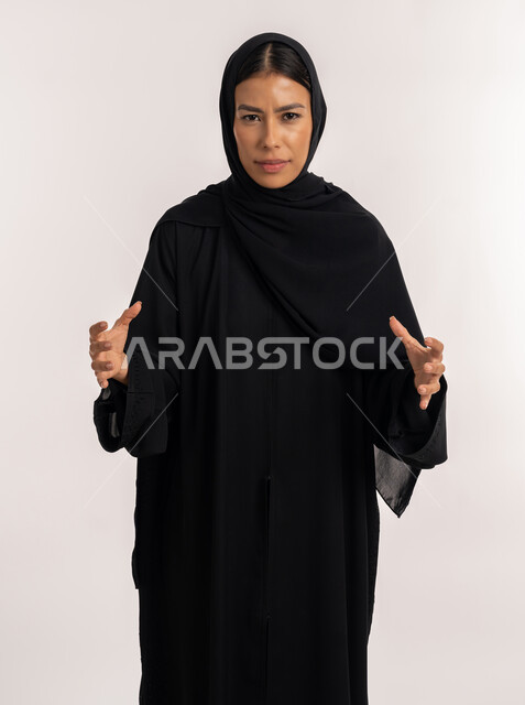 إيماءات الغضب و الانزعاج, بورتريه لامرأة عربية خليجية إماراتية ترتدي العباءة السوداء تنظر إلى الكاميرا بتعابير العصبية و النقاش بانفعال, شابة سعودية تمسك شيء ما, خلفية رمادية