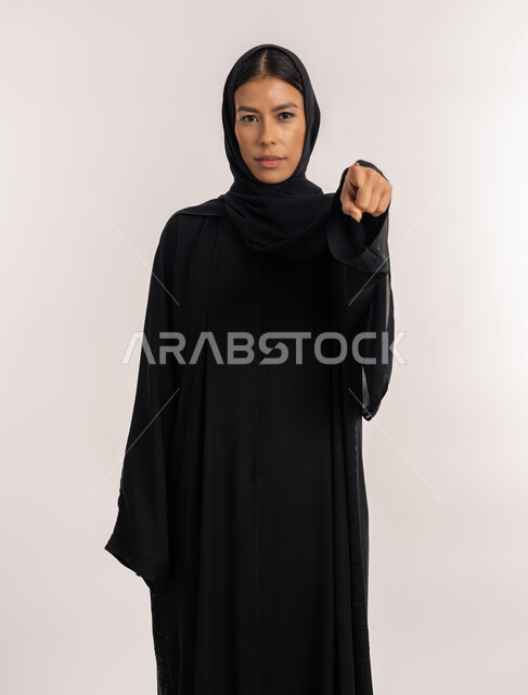 تعابير الجدية و الحزم, الاتهام و إلقاء اللوم, الإشارة إلى شيء ما, بورتريه لامرأة عربية خليجية إماراتية ترتدي العباءة السوداء تنظر إلى الكاميرا, شابة سعودية تشير بإصبع السبابة إلى الأمام, خلفية بيضاء