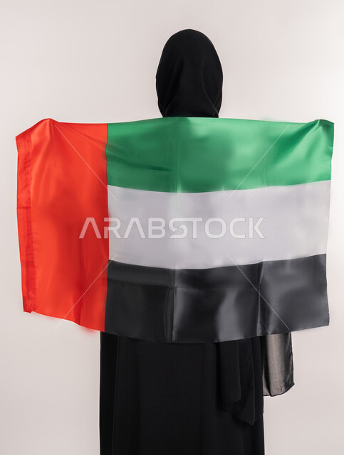 الاحتفال باليوم الوطني الإماراتي , الأعياد و المناسبات الوطنية, مفهوم الحب و الانتماء للبلاد, بورتريه من الخلف لامرأة عربية خليجية إماراتية ترتدي العباءة السوداء تحمل علم الإمارات العربية المتحدة على ظهرها, خلفية بيضاء