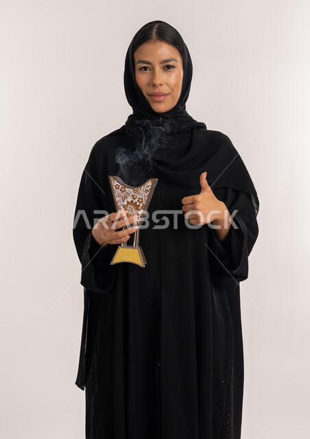 التطيب و التعطر باستخدام خشب العود و البخور, تعابير الرضا و الإعجاب, أصالة العادات و التقاليد الخليجية, بورتريه لامرأة مبتسمة عربية خليجية إماراتية ترتدي العباءة السوداء تمسك بيدها المبخرة التراثية المزخرفة و تنظر للكاميرا بإيماءات السعادة و السرور, شابة سعودية تشير بإصبع الإبهام للأعلى, خلفية بيضاء
