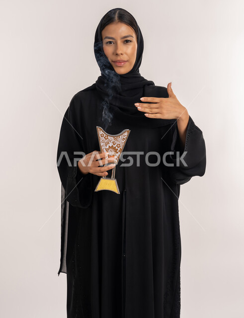 أصالة العادات و التقاليد الخليجية, مفهوم التطيب باستخدام خشب العود, النظر إلى الكاميرا بإيماءات السعادة و السرور, بورتريه لامرأة مبتسمة عربية خليجية إماراتية محجبة ترتدي العباءة السوداء تحمل بيدها المبخرة التراثية المزخرفة, شابة سعودية تقوم بالتعطر من حامل البخور, خلفية بيضاء