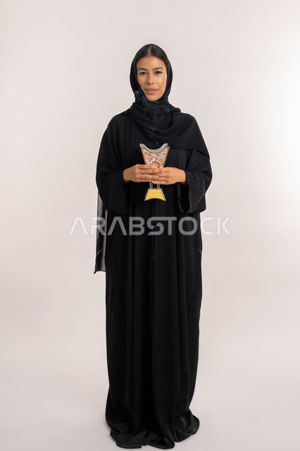 النظر إلى الكاميرا بإيماءات السعادة و السرور, مفهوم التطيب باستخدام خشب العود, أصالة العادات و التقاليد الخليجية, بورتريه لامرأة مبتسمة عربية خليجية إماراتية محجبة ترتدي العباءة السوداء تحمل بيدها المبخرة التراثية المزخرفة, شابة سعودية تقوم بالتعطر من حامل البخور, صورة للجسم بالطول الكامل, خلفية بيضاء