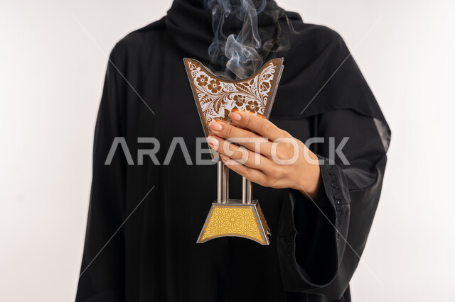 The concept of perfuming and perfuming, the use of agarwood on holidays and occasions, a close-up portrait of a veiled Arab Gulf Emirati woman wearing a black abaya, holding an ornate traditional incense burner in her hand, a young Saudi woman perfuming herself with incense, white background